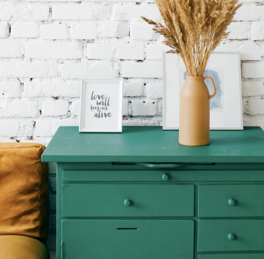 A dark green cabinet with three drawers against a white brick wall. On top of the cabinet, a picture with the phrase 'love will keep us alive', a beige vase with dried wheat and another picture leaning against the wall. Next to the cabinet, a yellow cushion.